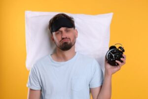 Unhappy sleep deprivation patient with pillow, alarm clock and sleep mask on yellow background