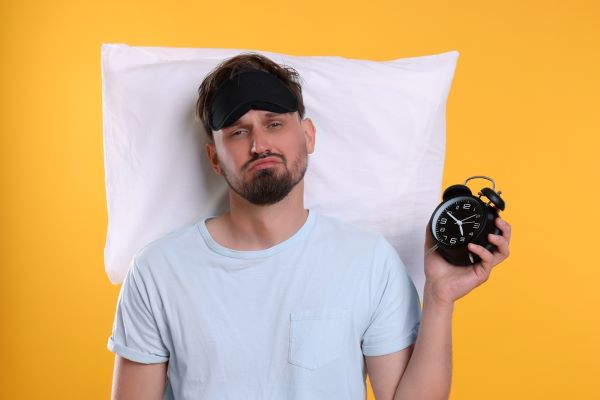 Unhappy sleep deprivation patient with pillow, alarm clock and sleep mask on yellow background